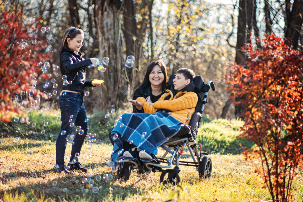 Mom with daughter and son with special needs outdoors. Mom is crouching close to son's wheelchair and daughter is blowing bubbles.

