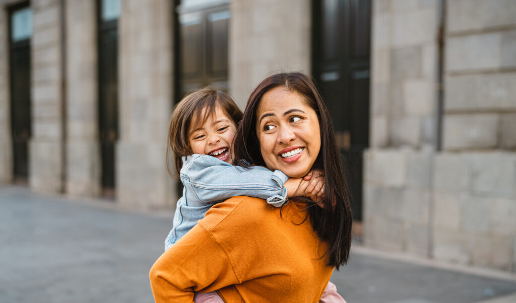 Daughter smiling ecstatically as mom carries her on her back, smiling and looking toward her leaving the law office after updating their asset plan to include a trusted guardian for their child. 
